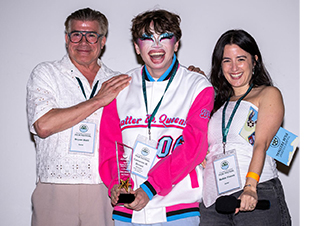 Three smiling people, one in drag makeup and colorful jacket holds an award. recipient