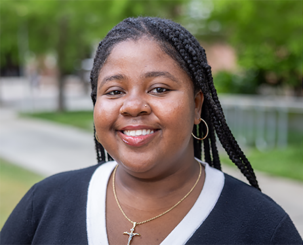 Smiling Black woman with braided hair and a gold cross necklace outdoors.