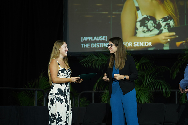 Economics student in floral dress receives senior award from professor on stage at ceremony.