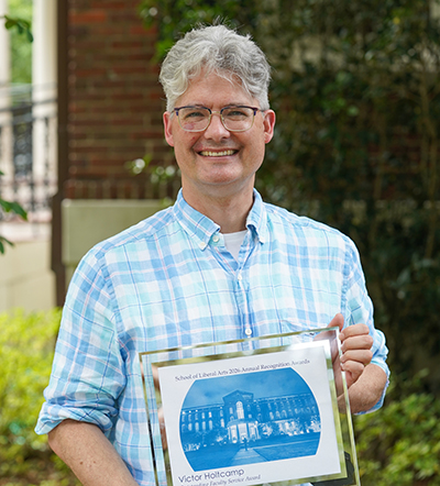 Smiling man in blue plaid shirt holds a framed award featuring a building graphic.
