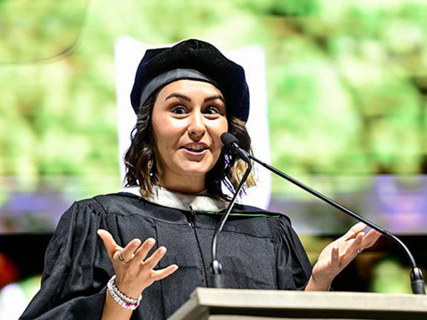 Smiling alumni speaker in cap and gown speaks animatedly at a podium with a microphone.