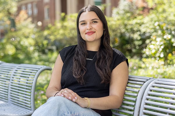 Young woman in black top and jeans smiles while sitting on a bench outside, campus background.