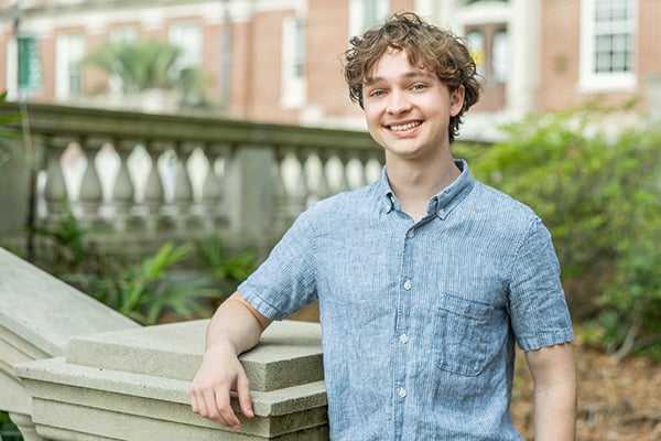 Smiling young man in a blue shirt leaning on a stone railing, building in background.