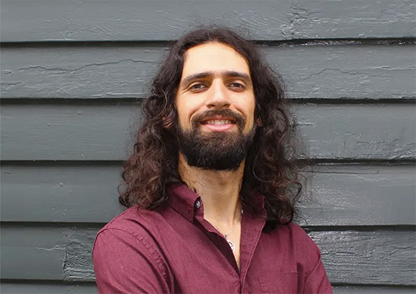 Smiling man with long curly hair and beard, wearing a maroon shirt against a gray wall.