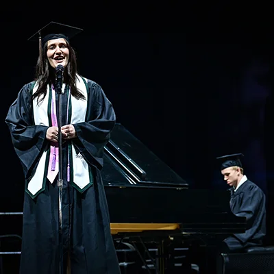 Graduate singing into a microphone, another plays piano on a dark stage.