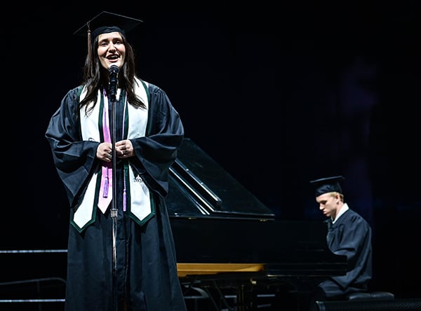 Graduates in cap and gown perform on stage; one sings, another plays piano.