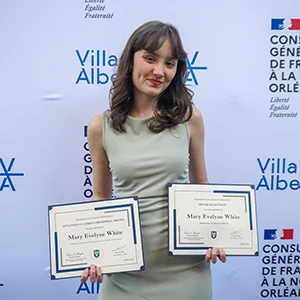 Mary Evelyne White holds two certificates against a French consulate banner.