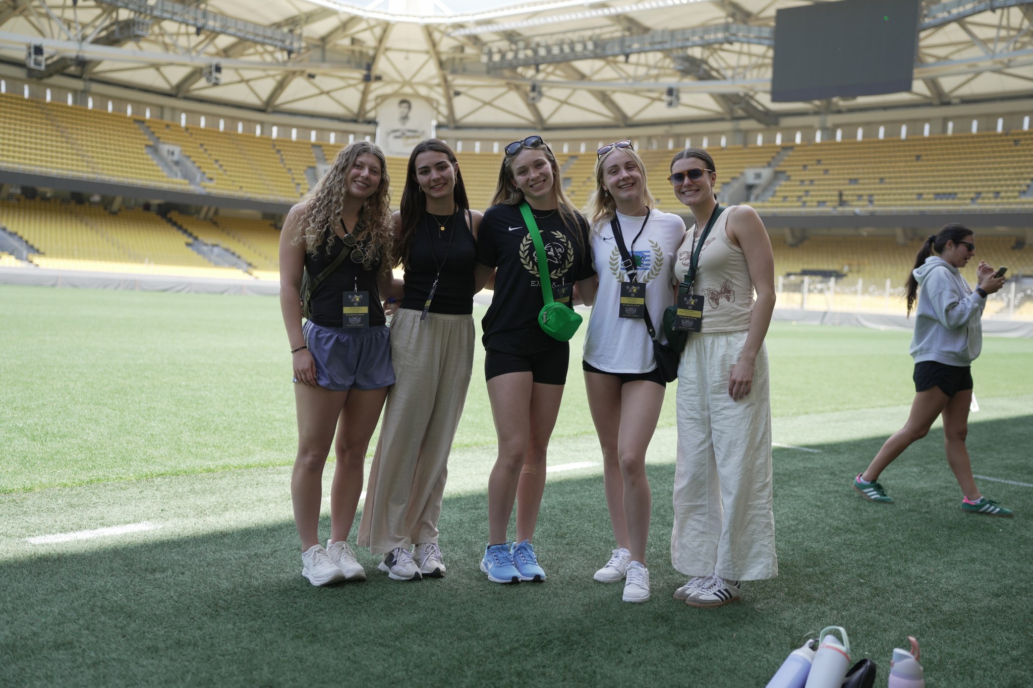 Five smiling young women stand on a green sports field inside a large stadium.