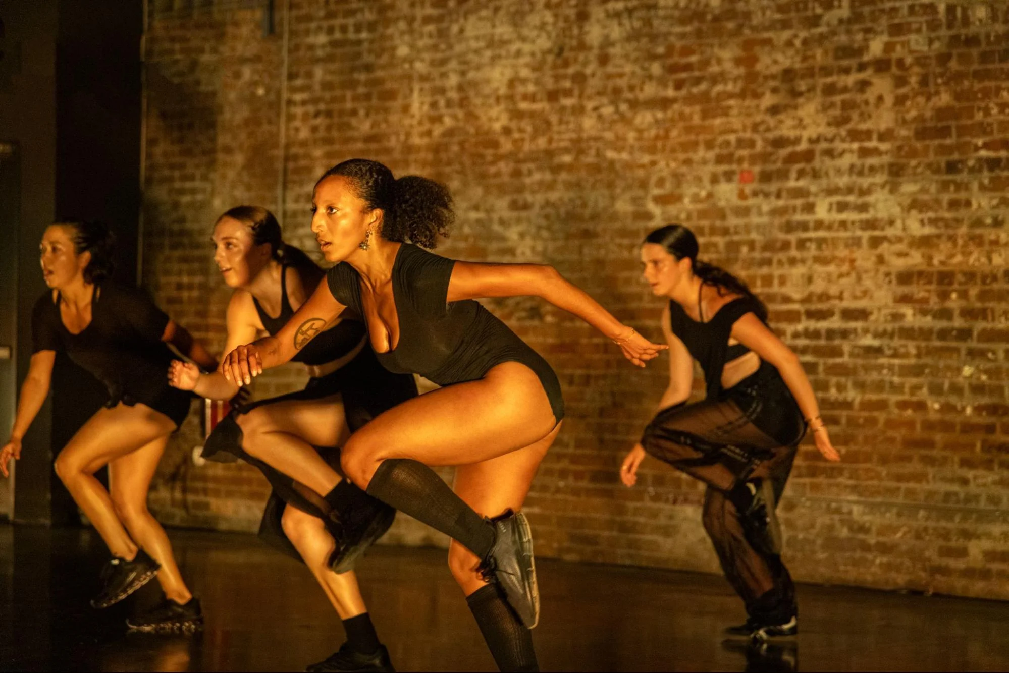 AT DAWN. Four dancers in black actively moving on stage against a brick wall at IDFNOLA 2026
