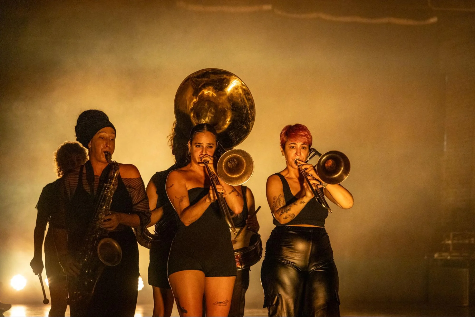 AT DAWN. Five women in black perform with brass instruments on a warmly lit, hazy stage.