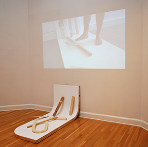 installation view of floor sculpture with video projection above it of person standing on sculpture