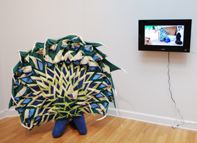 sculpture of figure in stuffed jeans and fabric peacock plumage, with video of figure dressed similarly in performance