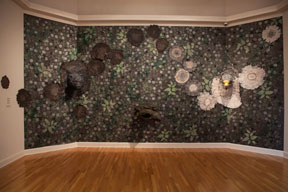 installation view of three adjacent walls in galley covered in dark foliate wallpaper with crane head and turtle head sculptures