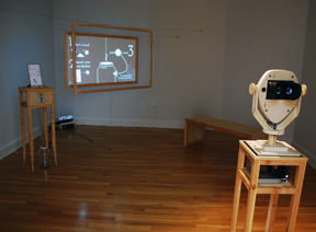 gallery room with virtual reality viewer, screen with video projection and small machine that takes coins