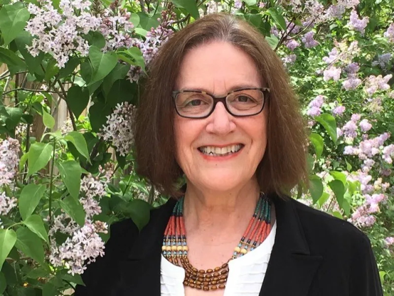 Teresa Toulouse in a Black Blazer and White Shirt in Front of a Bush with Light Pink Flowers 