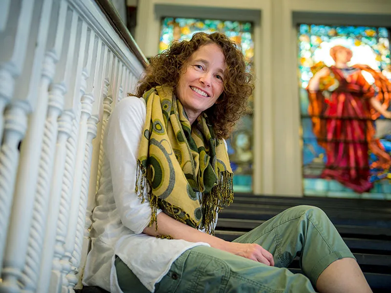 Dr. Toni Weiss sits comfortably on a set of stairs smiling down at the camera. She's wearing a 3 quarter length, white sleave shirt, green pants, and patterned yellow scarf. Light filters through large stained glass windows behind her.